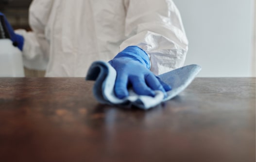 A person wearing blue gloves and a protective white suit cleans a surface with a cloth, emphasizing hygiene and cleanliness.